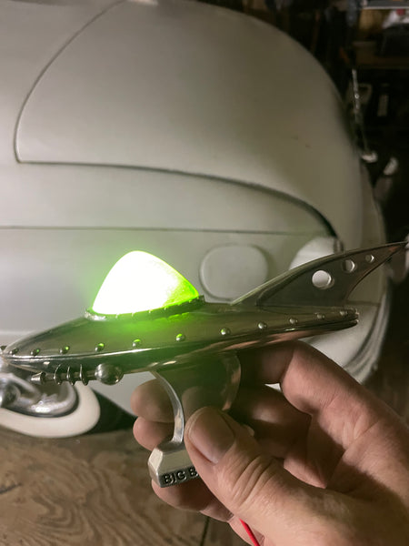 A hand holding a chrome silver retro-futuristic spaceship-shaped ring with a glowing green illuminated dome on top, set against a background of a white vintage car and workshop floor